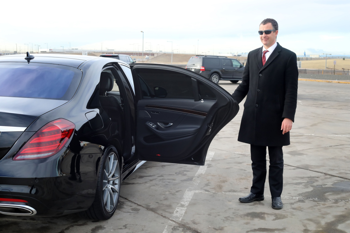A limo driver with sunglasses holding a black car door open for a passenger.