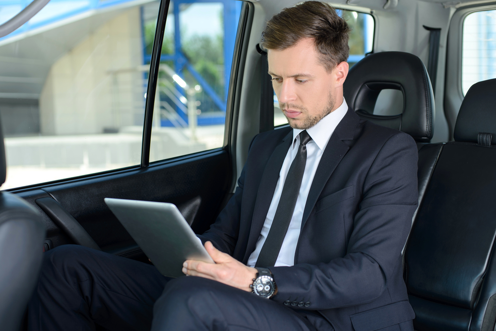 A man in a suit sitting in the back of a car and looking at a tablet.